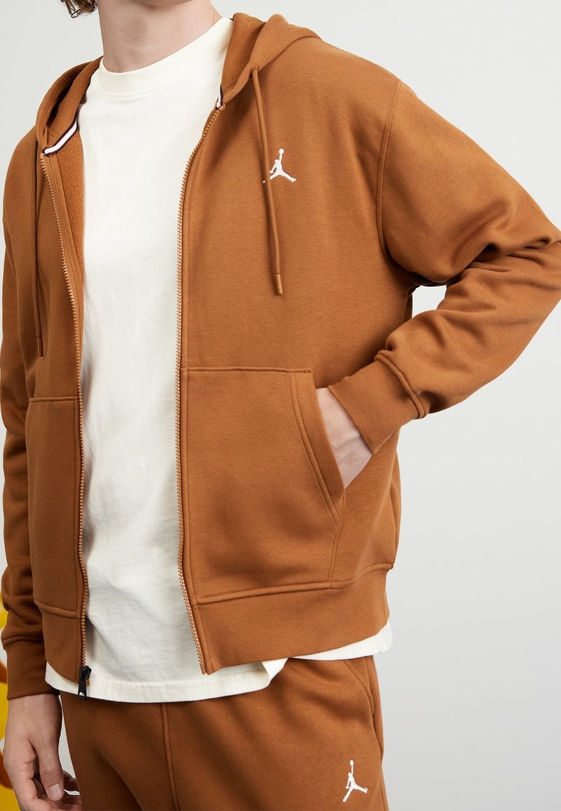 Personne portant un sweat à capuche marron zippé et un pantalon assorti avec le logo Jumpman blanc, main dans la poche, par-dessus un t-shirt blanc.