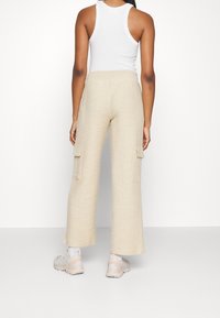 Osoba ubrana w kremowe, teksturowane spodnie cargo z szerokimi nogawkami, biały top na ramiączkach, białe skarpetki i beżowe sneakersy, stojąca na tle jednolitego białego tła.