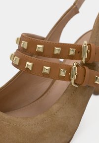 Καστόρινες γόβες σε ταμπά χρώμα με δύο λουράκια που φέρουν χρυσά πυραμιδοειδή καρφιά και λείες μεταλλικές λεπτομέρειες. Στρογγυλερή μύτη και πάτος με επένδυση για άνεση.