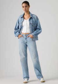 Γυναίκα που στέκεται με τα χέρια στις τσέπες, φοράει ανοιχτό μπλε τζιν μπουφάν και τζιν παντελόνι, λευκό κοντό τοπ με ρίγες και λευκά αθλητικά παπούτσια με μπλε και κόκκινες ρίγες.