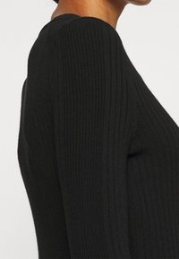 Haut noir à manches longues côtelé, présente une coupe ajustée, une surface texturée et un col rond classique, mettant en valeur une silhouette élancée.