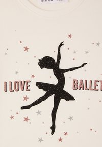Bawełniana koszulka T-shirt o kremowym tle, z czarną sylwetką tancerki baletowej oraz błyszczącymi różowymi i srebrnymi gwiazdkami i napisem „KOCHAM BALET.”