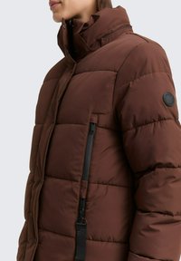 Braune Daunenjacke mit hohem Kragen, seitlichem Reißverschlussfach und einem runden Logo-Patch am Ärmel; aus gestepptem, strukturiertem Material.