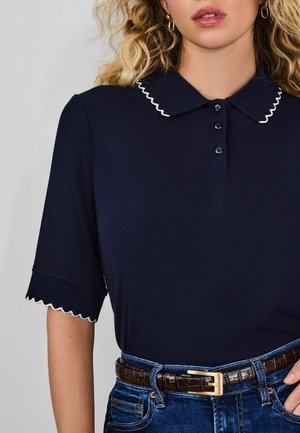 Woman wearing navy polo shirt with white scalloped trim on collar and sleeves, tucked into blue jeans with brown textured belt.