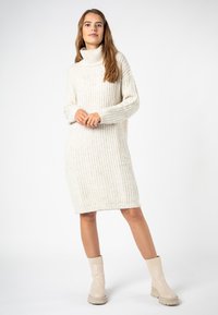 Cremfarbendes, geripptes Rollkragenkleid mit langen Ärmeln, knielanger Schnitt, kombiniert mit beigefarbenen Ankle-Boots mit dicker Sohle.