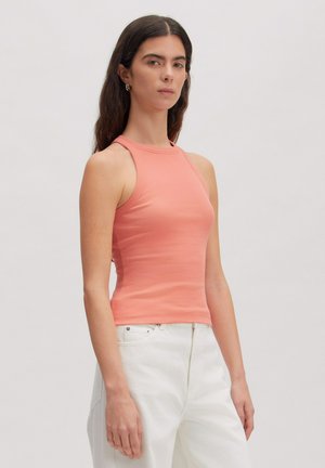 Frau mit langen dunklen Haaren trägt ein korallenfarbenes ärmelloses geripptes Top und weiße hoch taillierte Hosen, steht vor einem schlichten hellen Hintergrund.