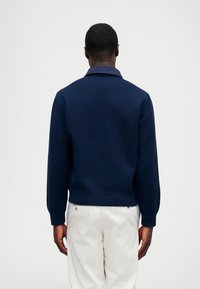 Σκούρο μπλε πλεκτό πουλόβερ με κολάρο, ριμπ μανίκια και ζώνη, σε συνδυασμό με ανοιχτόχρωμα παντελόνια.