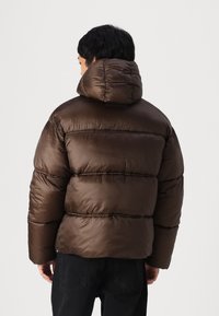 Chaqueta acolchada marrón con diseño de rombos, capucha y textura suave. Presenta un acabado ligeramente brillante y puños elásticos.