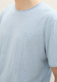 Camiseta de cuello redondo color azul claro, hecha de una tela suave, con un sutil logotipo grabado en el pecho y mangas cortas.