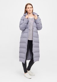 Langer, leichter Puffermantel in Hellgrau, mit Kapuze, Frontreißverschluss und horizontalen gesteppten Bereichen, getragen über einem gestreiften Shirt.