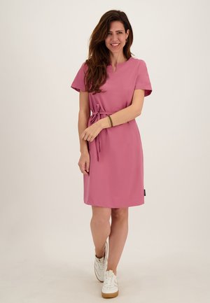 Vrouw in een knielange stofroze jurk met een tailleceintuur, witte sneakers en armband, staand glimlachend tegen een effen achtergrond.