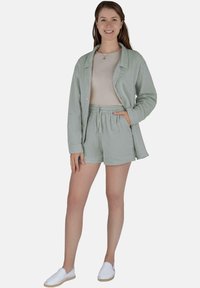 Frau in einem hellgrünen Leinenblazer und Shorts mit beigem Oberteil und weißen Slip-On-Schuhen, die mit einer Hand in der Tasche steht und lächelt.
