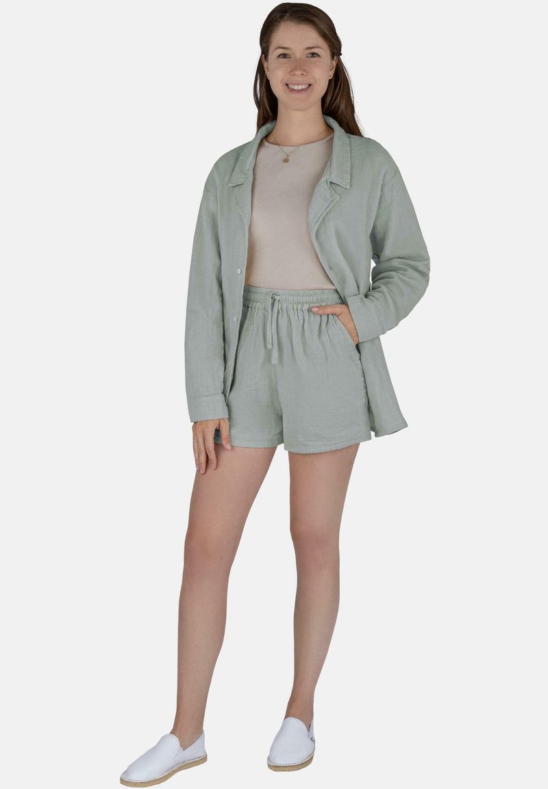 Frau in einem hellgrünen Leinenblazer und Shorts mit beigem Oberteil und weißen Slip-On-Schuhen, die mit einer Hand in der Tasche steht und lächelt.