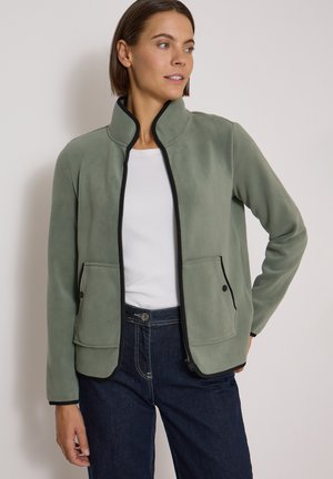 Femme portant une veste polaire zippée vert clair avec une bordure noire, chemise blanche et jean bleu foncé, debout avec une main sur la hanche.