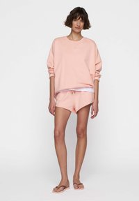 Mujer de pie con sudadera rosa clara y pantalones cortos a juego, con chanclas marrones, frente a un fondo blanco liso.