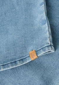Lichtblauwe denimstof met zichtbare diagonale weving en een klein bruin leren label dicht bij de zoom genaaid.
