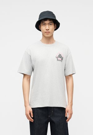 Fiatal férfi világosszürke pólóban, melynek mellrészén egy kis rózsaszín és fekete motívum látható, fekete vödörkalappal, egyszínű háttér előtt állva.