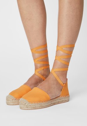 Espadrilles orange avec une tige en toile, semelle en corde de jute et des brides en tissu croisées autour des chevilles. Texture lisse, design à dos ouvert.