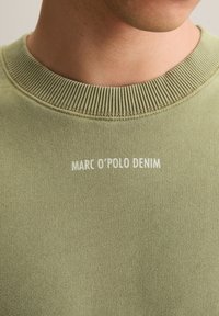 Close-up van een persoon die een lichtgroene sweater met ronde hals draagt, met witte tekst "MARC O'POLO DENIM" op de borst.