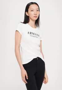 Balts īsām piedurkņu T-krekls ar melnu "Armani Exchange" logotipu, saskaņots ar melniem pieguļošiem biksēm, attēlots uz neitrāla fona.