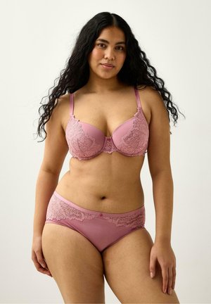 Kvinde med langt krøllet hår iført pink blondesexet bh og matchende trusser, stående mod en ensfarvet lys baggrund, vendt mod fronten.