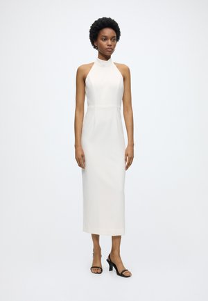 Frau mit kurzen lockigen Haaren, die ein ärmelloses, weißes Midi-Kleid mit hohem Kragen und schwarze offene High Heels trägt, steht vor einem einfarbigen weißen Hintergrund.