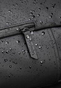 Black waterproof bag with a smooth texture, featuring droplets of water and a silver zipper pull with a tag attached.