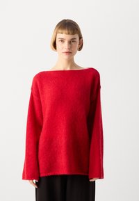 Rød oversized genser med vide skulderlinjer, lange ermer og en myk, teksturert finish. Bært over mørke bukser, minimalistisk design, uten mønstre.