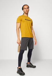 Gul kortärmad T-shirt med svart "Mammut"-logga, kombinerad med mörkgrå shorts och marinblå leggings, samt svarta träningsskor. Håller en beige vattenflaska.