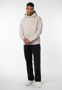 Light beige hoodie with front pocket and orange logo, paired with black jeans and white sneakers. Smooth texture, casual fit.