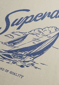 Croquis bleu d'un voilier sur l'eau avec des nuages au-dessus et le mot "Superdry" en cursive sur un tissu beige.