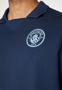 Polo simplu de culoare bleumarin, cu guler, având logo-ul embrodat al echipei Manchester City, de culoare bleumarin deschis, pe piept. Material din bumbac moale, cu o textură netedă.