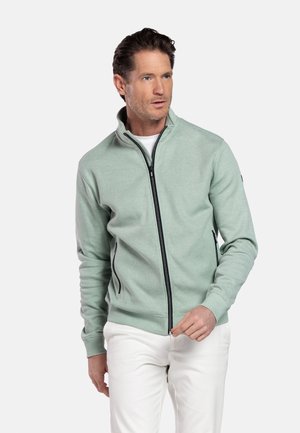 Man draagt lichtgroene jas met ritssluiting over wit shirt en witte broek, staand tegen een eenvoudige lichte achtergrond.