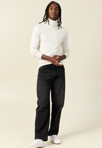 Suéter de cuello alto crema con puños y dobladillo de canalé, combinado con vaqueros negros holgados y zapatillas blancas, centrado sobre un fondo claro.