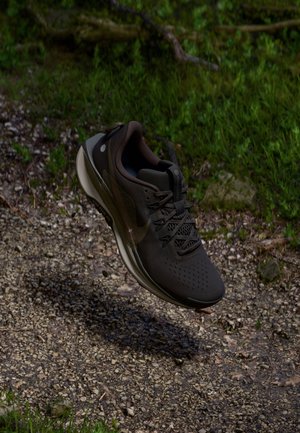 Chaussure de sport noire avec des lacets flottant au-dessus d'un sentier rocailleux entouré d'herbe verte et de feuillage.