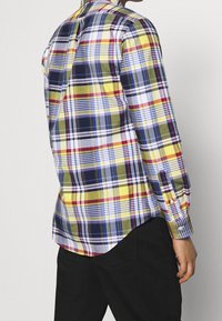 Rutig skjorta med gula, blå, gröna och röda färger, långa ärmar, knapputförande krage och rundad nederkant. Tillverkad av lätt material.