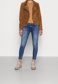 Chaqueta biker de ante marrón, top ajustado color canela, vaqueros pitillo azules y zapatillas beige. La chaqueta presenta cremalleras y una superficie texturizada.