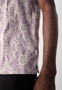 Gros plan d'une chemise violette avec une grande broderie florale blanche sur un bras à la peau foncée, montrant la texture du tissu et les détails du motif.