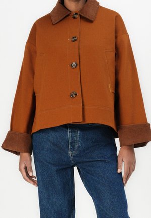 Personne portant une veste brune boutonnée avec manches larges et col, associée à un jean bleu, debout devant un fond uni clair.