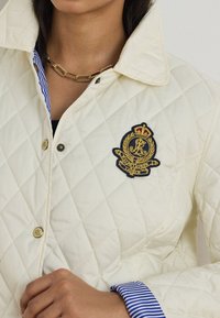 Femme portant une veste matelassée crème avec un écusson Ralph Lauren et des boutons dorés, sur une chemise rayée bleu et blanc.