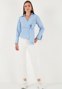 Donna in piedi che indossa una blusa avvolgente a righe blu, jeans bianchi, sneakers bianche, orecchini a cerchio d'oro e collane d'oro a strati su uno sfondo bianco.