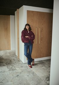 Veste bomber bordeaux, haut blanc, jeans bleus et ballerines rouges. L'arrière-plan présente des meubles en bois clair et un sol en pierre grise.