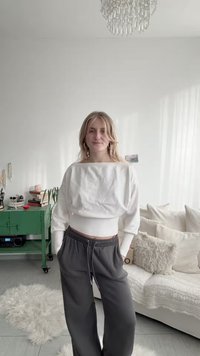 Jeune femme en pull court blanc et pantalon de jogging foncé, se tenant dans un salon lumineux avec une armoire verte, un canapé blanc et un tapis moelleux.