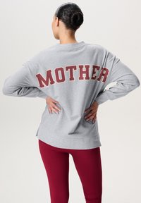 Szara bluza z długim rękawem i luźnym fasonem, z czerwonym napisem "MOTHER" na plecach, w zestawie z czerwonymi legginsami. Gładka tekstura, swobodny styl.