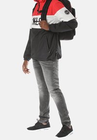 Persona che indossa una giacca rossa, bianca e nera con uno zaino, jeans slim grigi e scarpe da ginnastica nere, in piedi su uno sfondo bianco.