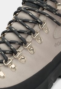 Bota de caminhada bege com um cabedal em couro liso, sola de borracha preta, ilhós de metal e atacadores escuros com padrão entrelaçados pelo cano.