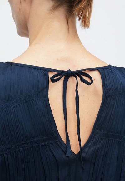 Partea superioară a spatelui unei femei, purtând o bluză plisată albastru închis, cu decupaj în formă de V în spate, legat cu un papion la gât.