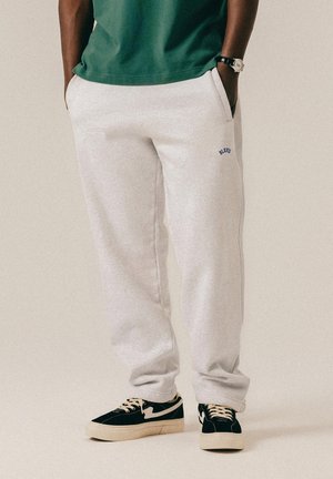 Persona che indossa pantaloni della tuta grigio chiaro con logo blu "BLKTVS", maglietta verde, sneakers nere e bianche, in piedi con le mani nelle tasche.
