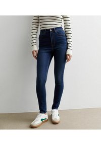 Vaqueros de mezclilla azul oscuro de cintura alta con una textura suave, que cuentan con cinco bolsillos y costuras en contraste. Combinados con zapatillas blancas.