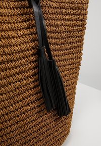 Brązowa pleciona torba o teksturowanej powierzchni, ozdobiona czarnymi skórzanymi frędzlami jako akcent. Okrągły kształt, widoczna solidna konstrukcja.
